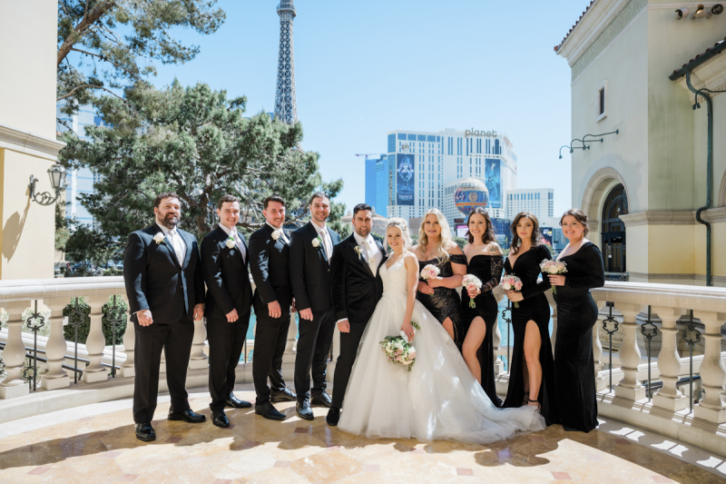 A wedding party lines up and poses for a photograph. They are standing on a balcony at the Bellagio Hotel and Casino which is overlooking the Las Vegas Strip. The four groomsmen are on the left and wear black suits. The groom and bride are in the middle. The four bridesmaids are lined up on the right and wear black dresses.