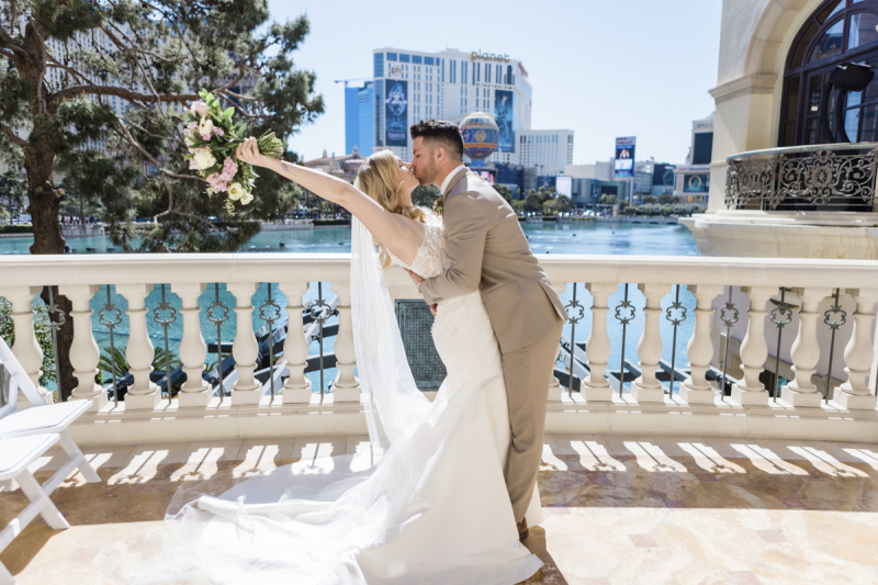 A bride tips slightly backwards and raises her flowers as her new husband kisses her on a balcony at the Bellagio Hotel and Casino on the Las Vegas Strip. She is wearing a long white dress with a long vail. He is wearing a light brown suit. Behind them is the Bellagio's Lake and the Paris Las Vegas Hotel and Casino.