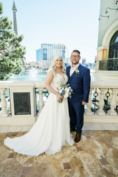 A bride and groom stand side-by-side together and pose for wedding pictures on a balcony overlooking the lake in front of the Bellagio Hotel and Casino in Las Vegas with the Paris Las Vegas's Eiffel Tower in the background. She wears a long sleeveless dress. He wears a blue suit with an unbuttoned dress shirt.