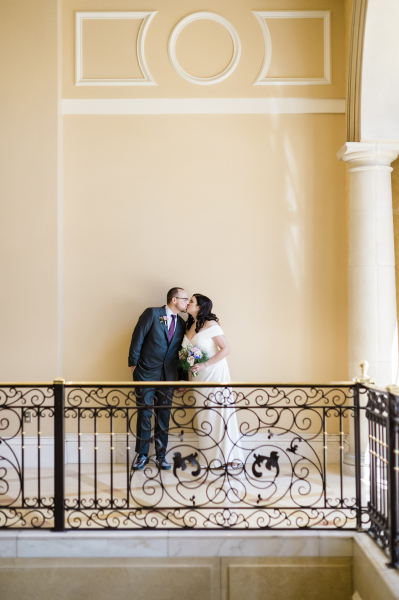 A groom and bride are kissing on the far side of an interior courtyard balcony. The Italianate architecture is highlighted by geometrical wall moldings on the wall behind them, a Tuscan column on the right edge of the picture, a brass and black railing in front of them and a white marble floor.