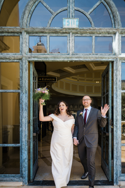 A bride and groom hold hands and walk through arched French doors with their arms raised in celebration on their wedding day. A blue sky is reflected in the uppermost windows above the door.