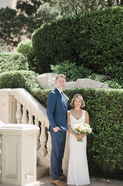 A bride and groom stand at the base of a short Italianate staircase in a garden. Green bushes rise behind them. The tall groom is wearing a blue suit. The much shorter bride is wearing a simple floor-length white dress.