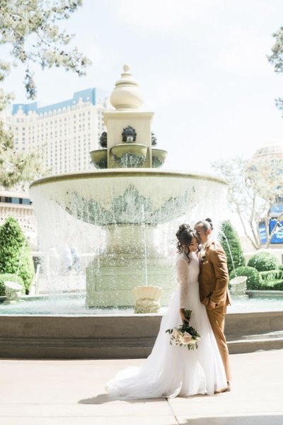 A bride and groom pose for wedding photos on a sunny day in front of an ornate water fountain at the Bellagio Hotel and Casino in Las Vegas. The groom appears to be whispering in the bride\'s left ear as she holds her flowers down on her right side.