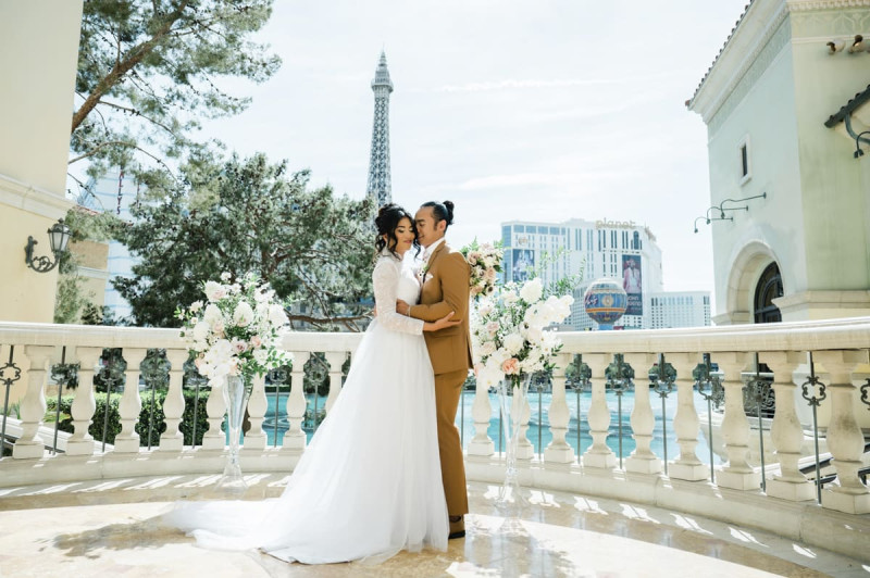 A bride and groom hold each other as the groom nuzzles into the side of his wife\'s head. They are standing on a balcony overlooking the lake in front of the Bellagio Hotel and Casino in Las Vegas with the Paris Las Vegas\'s Eiffel Tower in the background.