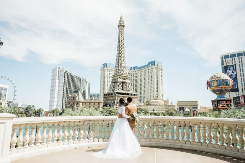 A bride and groom hold each other around the waist while looking out across the Las Vegas Strip. They are standing at a railing which shields the lake at the Bellagio Hotel and Casino in Las Vegas. Parts of The LINQ, the Flamingo, Bally\'s, Paris, and Planet Hollywood are all visible.