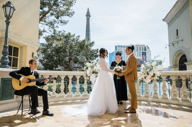 A wedding ceremony is being conducted by an officiant on a balcony at the Bellagio Hotel and Casino in Las Vegas. An acoustic guitar player sits and plays his instrument to the left of the couple while the ceremony takes place.