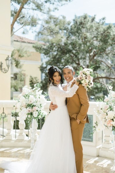 A bride and groom embrace and turn towards the camera with great big smiles. They are flanked by tall white floral displays as they pose for wedding day pictures on a balcony at the Bellagio Hotel and Casino in Las Vegas.