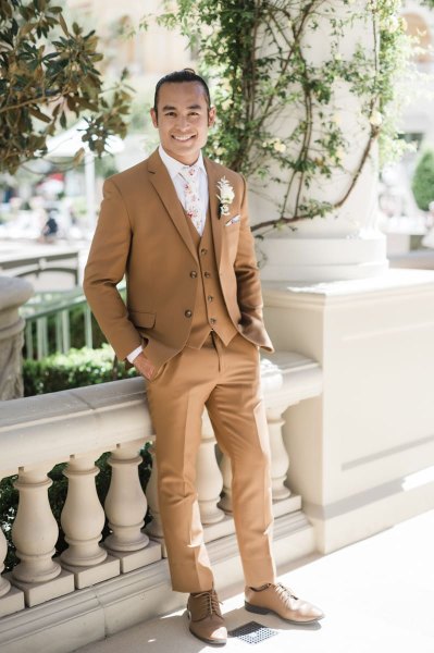A groom in a tan three-piece suit poses against a Romanesque cement railing. His hands rest in his pockets, and he has a smile. There are vines encircling the pillar behind him.