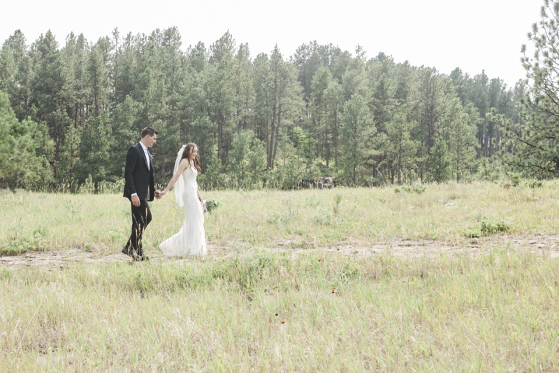 A bride leads a groom by the hand as they walk through a grassy meadow along a path that cuts through the grass. It is a sunny wedding day in the Black Hills of South Dakota.