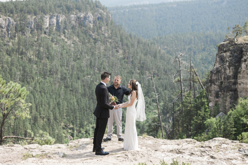 Wide shot of a wedding ceremony being conducted on the edge of a mountainous cliff in the Black Hills. Only the bride and groom and an officiant are present.