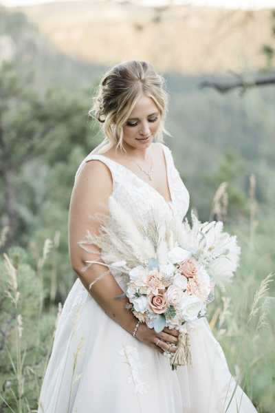 A portrait of a bride standing in a field of tall grass and admiring her bouquet of flowers.