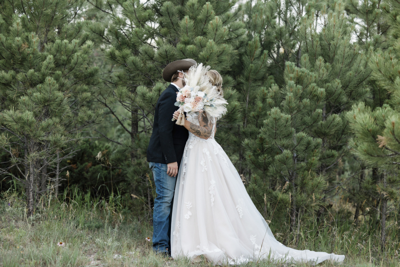 A groom and bride pose in front of a stand of pine trees. They are presumed to be kissing but the woman holds her flowers in front of their faces to block the camera.