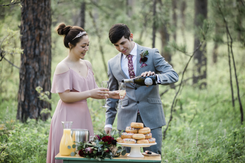 A bride holds a Champagne glass for her new husband to fill from a bottle. He pours as she watches. They are celebrating their wedding with mimosas and donuts in the woods of the Black Hills of South Dakota.