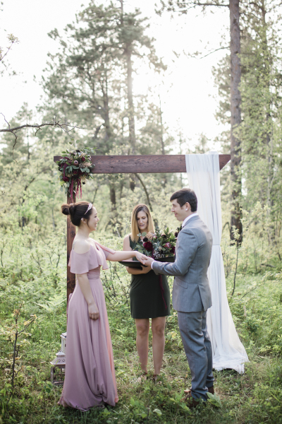 A bride and groom and their female officiant stand in front of a wooden arch during a wedding ceremony in the woods. The bride extends her hand and arm towards the groom as they hold hands.