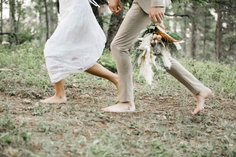 A closeup of a bride and groom running barefoot down a wooded path. Only their feet, ankles and legs are visible. A bouquet of flowers dangle from the groom\'s left hand and he holds the woman\'s hand in his right hand.