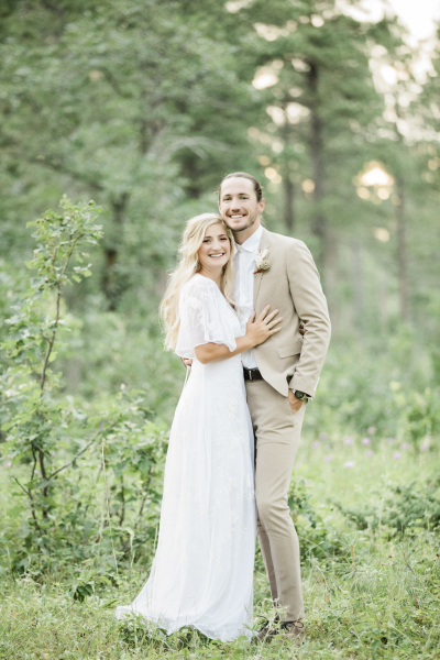 A vertically framed full body image of a bride and groom holding each other as they stand in the woods on their wedding day. They have just gotten married in the Black Hills of South Dakota.