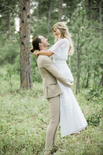 A groom in a tan suit lifts his new wife off the ground as they pose for wedding pictures in the Black Hills of South Dakota.