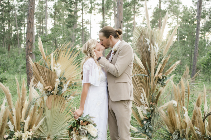 A bride and groom kiss in the woods. They are standing in front of a big floral display made up of dry palm leaves, pampas grass, and silver dollar eucalyptus.