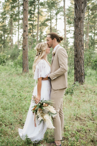 A bride and groom embrace in the woods of the Black Hills of South Dakota. He holds her at the waist and kisses her on the forehead while she holds a floral bouquet at her side.