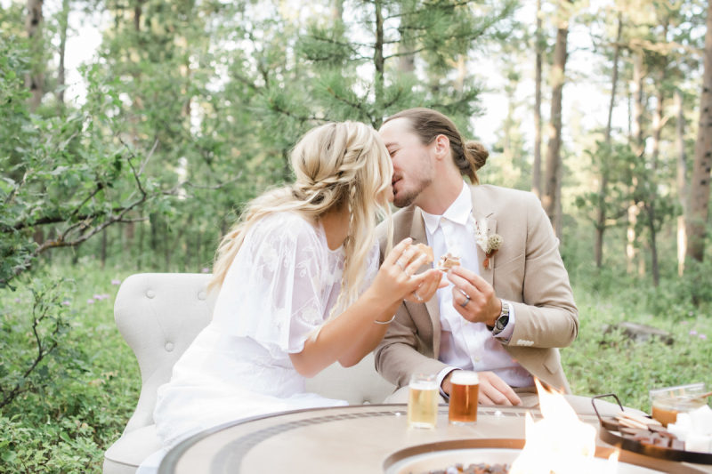 A bride and groom sit at a propane fire pit and share s\'mores and a kiss. There are also small glasses of beer on the edge of the fire pit.