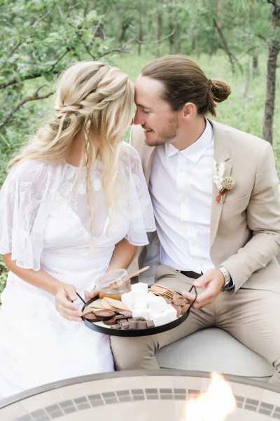 A bride and groom sit at a gas fire pit and nuzzle their noses as they hold a plate of s\'mores between them as part of their wedding day celebration in the Black Hills of South Dakota.