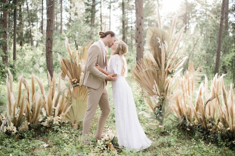 A bride and groom kiss in the woods. They are standing in front of a big floral display made up of dry palm leaves, pampas grass, and silver dollar eucalyptus. The sun is streaming in from the background between the trees.