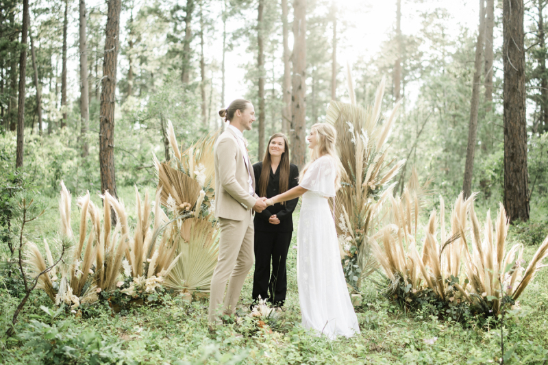 A bride and groom take part in their Black Hills South Dakota wedding ceremony in front of a big floral display made up of dry palm leaves, pampas grass, and silver dollar eucalyptus.
