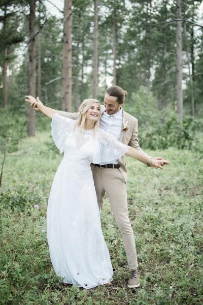 A bride and groom pose for a playful wedding photo in the woods. She stands in front of him and lifts her arms straight out at her sides while he holds her hands.
