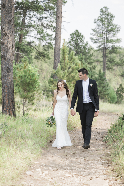 A young bride and groom walk hand-in-hand towards the camera on a dirt path through a pine tree forest. Their wedding is taking place in the Black Hills of South Dakota.