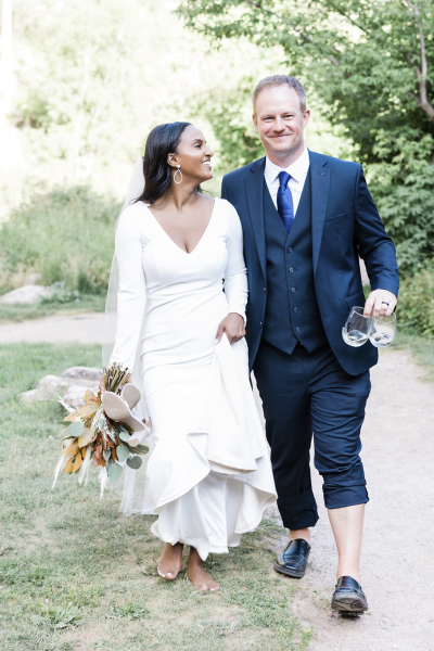 A mixed-race wedding couple walks down a path with wine glasses. She is barefoot and holds her shoes and flowers. He holds the glasses and has one pant leg rolled up to his shin.