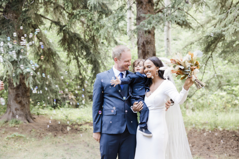 A couple poses for wedding photos in the woods with a young child. The woman holds the boy in one arm and her flowers in the other while the man stands closely at her side. Soap bubbles are drifting through the frame.