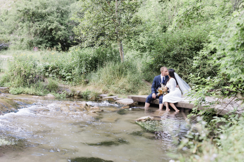 A groom and bride sit on the edge of a small footbridge above a mountain stream and dangle their bare feet into the water. They kiss as they pose for wedding photos in the Black Hills of South Dakota.
