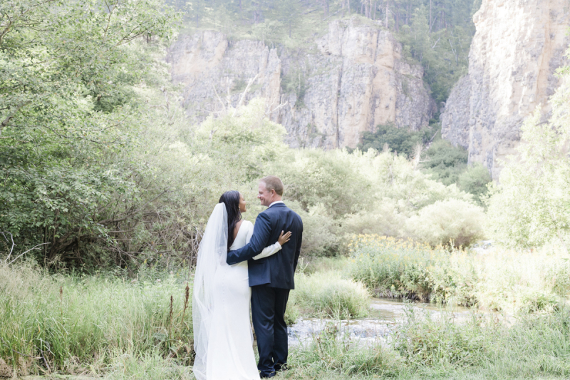 A bride and groom stand facing a mountain stream and turn their heads to smile at each other. They have their backs to the camera and their arms around each other.