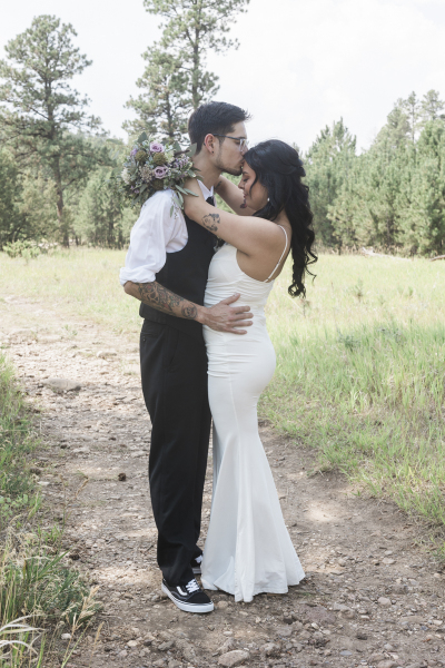 A groom and bride embrace as they pose for pictures on their wedding day. They are standing on a dirt path leading through a grassy meadow in the Black Hills of South Dakota.
