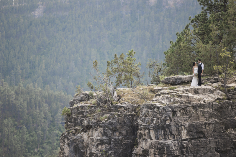 A long shot of a distant bride and groom standing on the other side of a cliff. They are posing for wedding pictures following their ceremony in South Dakota\'s Black Hills region.