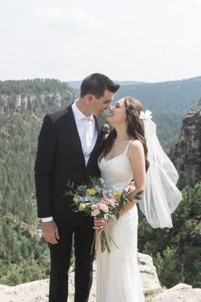 A young groom and bride are about to kiss as they stand near the edge of a rocky cliff overlooking a mountainous pine tree covered canyon on a sunny day.