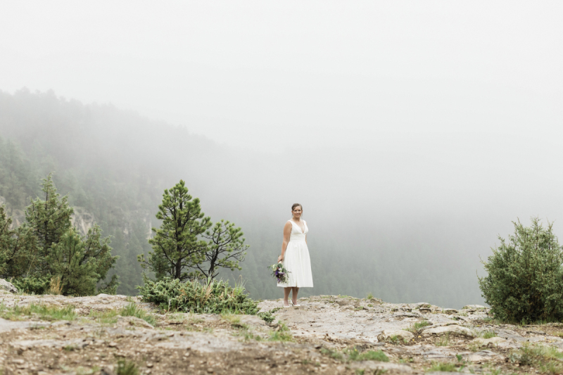 A wide shot of a bride in a simple white dress standing on a rocky ledge in the Black Hills on her wedding day. Fog fills the mountainous canyon behind here.