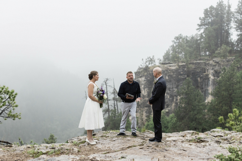 A bride and groom stand listening to their wedding officiant during their wedding ceremony which takes place near the edge of a mountainous cliff in the Black Hills.