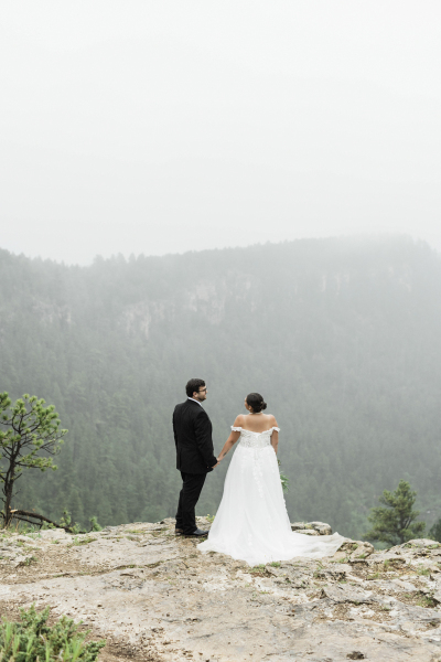 A wide shot of a groom and bride standing near the edge of a cliff. They are holding hands and facing each other while turning away from the camera. A wide shot of a groom and bride standing near the edge of a cliff. They are holding hands and facing each other while turning away from the camera.