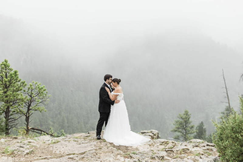 A wide shot of a couple embracing on their wedding day. They are posing for wedding photographs on a cliff overlooking a mountainous Black Hills South Dakota canyon on a foggy day.