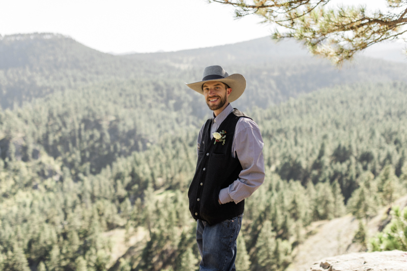 A groom in a cowboy hat stands under the shade of a pine tree and smiles for the camera. Behind him is a sunny pine tree covered mountain slope which is part of the Black Hills of South Dakota.