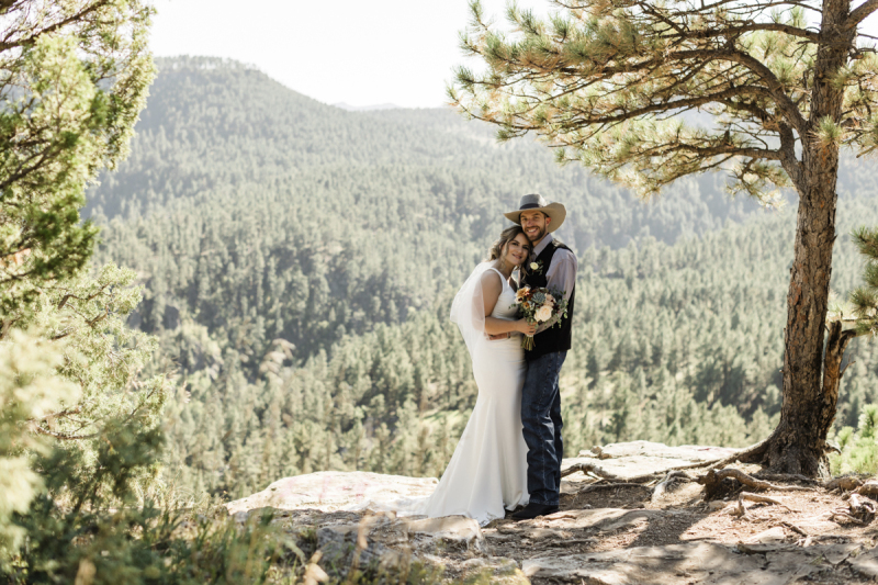 A bride and her groom hold each other closely at the waist and smile for the camera as they pose under a pine tree at the edge of a mountainous cliff in the Black Hills of South Dakota.