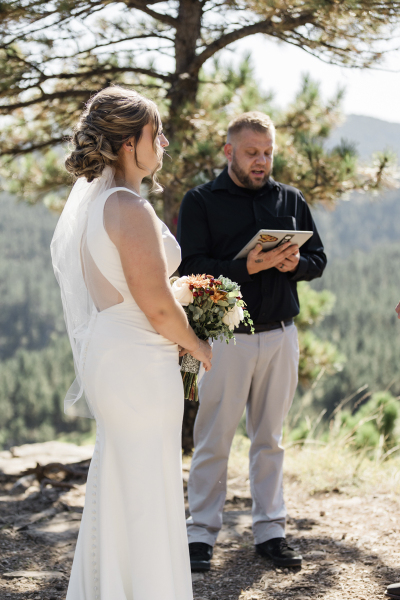 A South Dakota bride in a white long sleeveless dress stands on the left side of the frame listening to a wedding officiant who stands underneath a pine tree in the background. She is looking off camera to the right.