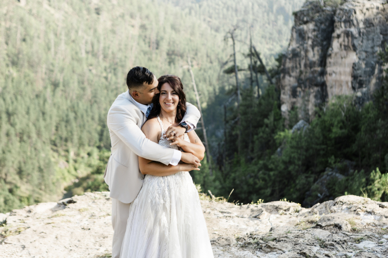 A happy looking couple poses for wedding photographs in front of a steep cliff that rises behind them. The groom leans over the bride\'s shoulders from behind and gives her a hug and kiss while she smiles at the camera.