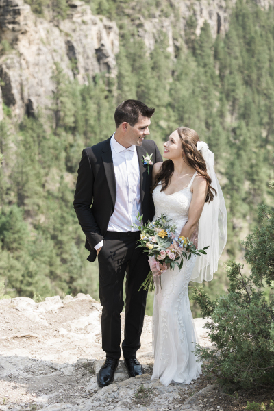 A young couple dressed in traditional wedding attire turn their heads to look at each other as they pose for pictures in the Black Hills of South Dakota on their wedding day.