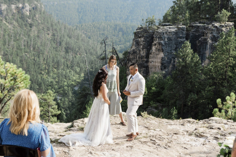 A wedding ceremony is taking place near the edge of a cliff. The groom appears to be holding a wedding ring which he is about to give to the bride. An officiant looks on between them and one guest in a blue dress is in the foreground.