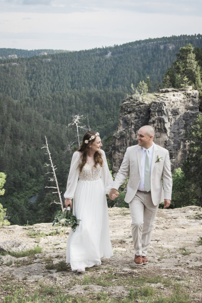 A bride with long brown hair and a floral crown holds her new husband\'s hand as the couple walks towards the camera as they pose for wedding photos in the Black Hills of South Dakota.