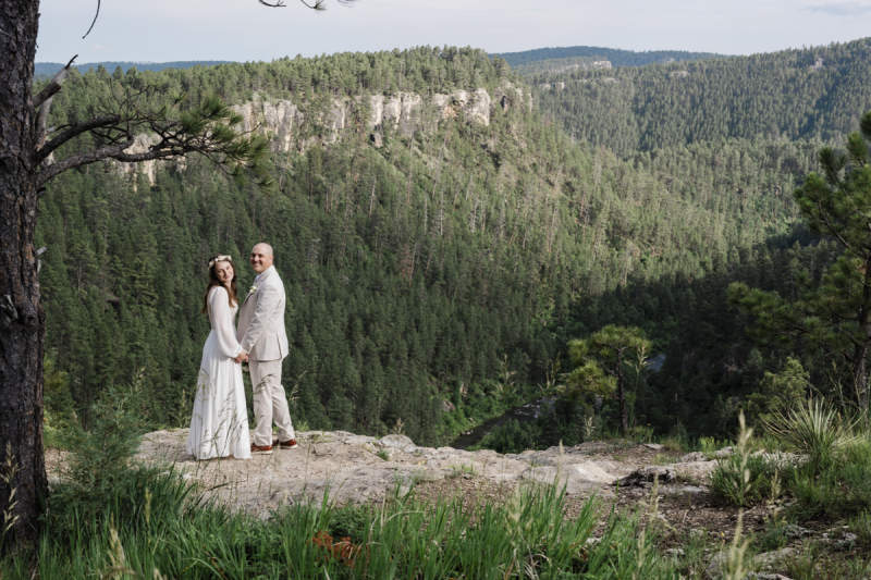 A pine tree covered mountain cliff fills the background as a couple is photographed in a medium shot holding hands and smiling. They have just been married in the Black Hills of South Dakota.