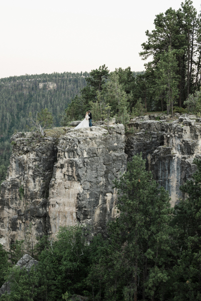 A bride and groom can be seen in the distance on a faraway cliff. The couple is posing for wedding photos following their ceremony in the Black Hills of South Dakota