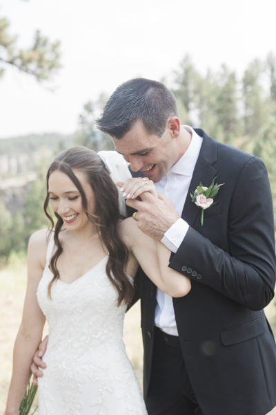 A medium shot from the waist up of a bride and groom laughing and holding hands on their wedding day. The groom stands behind the bride and lifts her left hand towards his lips.